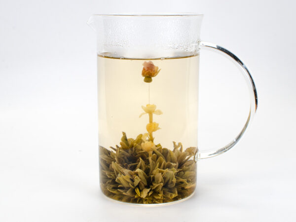 A full glass pitcher holding a fully bloomed ball of green tea with a chain of jasmine flowers topped with a single rose bud springing from the center.